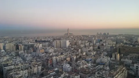 Casablanca buildings fill the foreground beneath a pale sunrise sky, with the Hassan II Mosque minaret by the Atlantic.