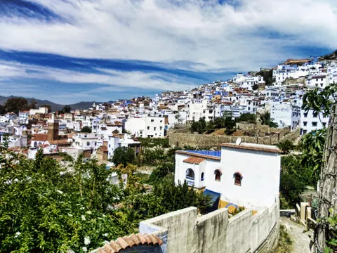 White and pale-blue buildings climb a hillside in Chefchaouen, with trees and an ochre tower below a cloudy sky.