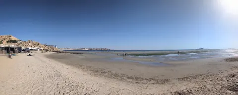 People stand beside shallow lagoon water at Dakhla’s PK25 beach, with sandy banks and beach shelters to the left.