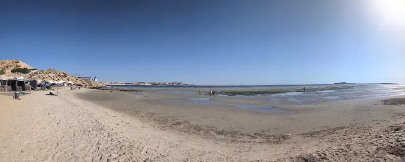 People stand beside shallow lagoon water at Dakhla’s PK25 beach, with sandy banks and beach shelters to the left.