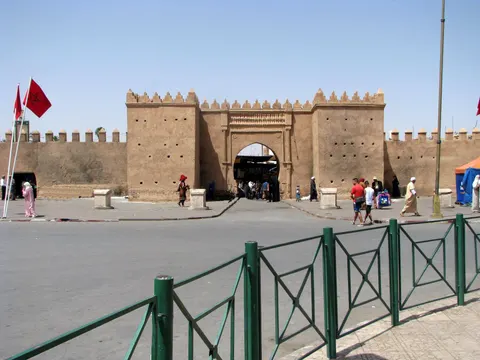 People pass through the ochre, crenellated Bab Sidi Abdelwahab gate in Oujda, with Moroccan flags beside the square.