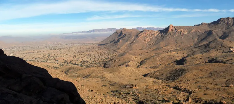 Rocky brown mountain ridges overlook a broad valley dotted with trees in the Tafraout region.