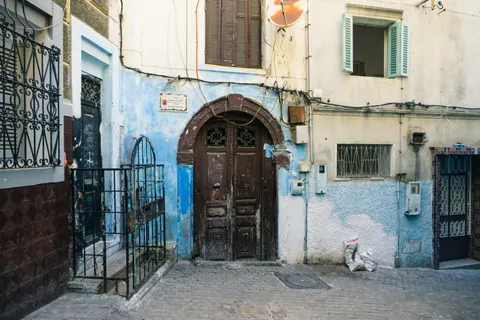 A dark wooden doorway in a weathered white and blue facade, with shutters and overhead wires in Tangier.